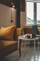 Cozy home interior with mustard armchair, burning candle in glass jar, and laptop on a small white table. Warm natural light and peaceful atmosphere