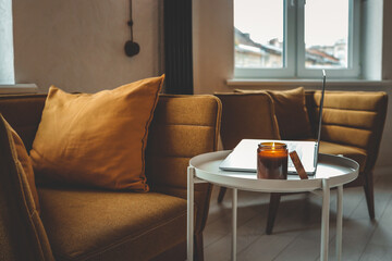 Cozy home interior with mustard armchair, burning candle in glass jar, and laptop on a small white table. Warm natural light and peaceful atmosphere
