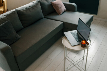 Burning candle and laptop on small white table in cozy loft interior with gray sofa, pillow, floor...