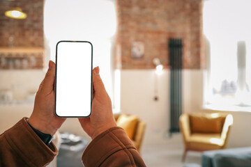 Person holding smartphone with blank screen in cozy loft interior with mustard armchairs, gray...