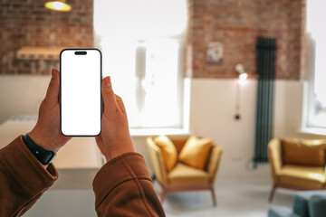 Person holding smartphone with blank screen in cozy loft interior with mustard armchairs, gray...