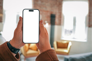 Person holding smartphone with blank screen in cozy loft interior with mustard armchairs, gray...