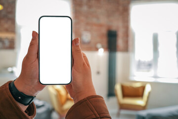 Person holding smartphone with blank screen in cozy loft interior with mustard armchairs, gray...