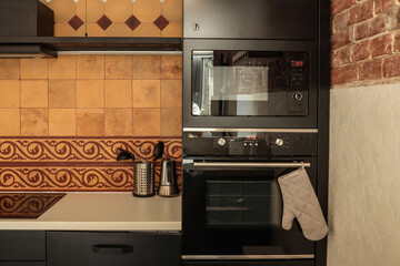 Modern kitchen with black appliances, beige and terracotta tiles, and vintage ceramic pattern. Oven with mitt, utensils, and coffee pot on clean countertop