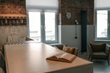 Cozy interior with an open book placed on a light kitchen counter, soft natural light from windows, modern loft-style design with brick walls and leather armchairs