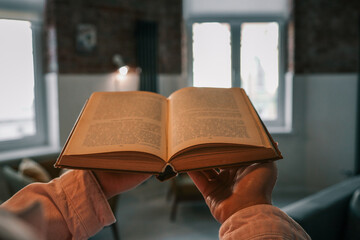 Close-up of an open vintage book held in hands, with a cozy modern living room in soft focus in the...