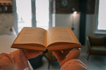 Close-up of an open vintage book held in hands, with a cozy modern living room in soft focus in the...