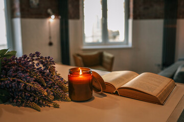 Lit candle, purple flowers, and an open vintage book on a light wooden table in a cozy loft-style...