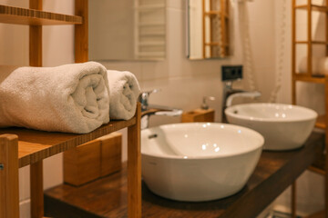 Close-up of rolled white towels on wooden shelf next to modern ceramic sinks and chrome faucets in...