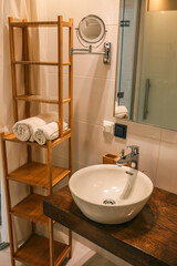 Modern bathroom interior with a single white ceramic washbasin, wooden shelf unit, and mirror on tiled wall
