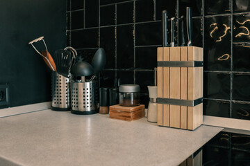 Modern kitchen countertop with black utensils in metal holders, wooden knife block, and spice jars...