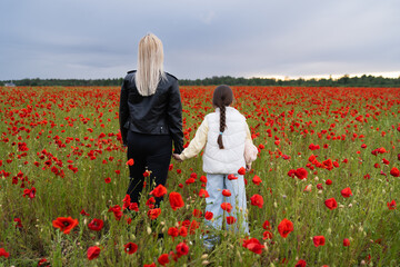 Mother and daughter, holding hands, walk through a bright field with red poppies under a gloomy,...