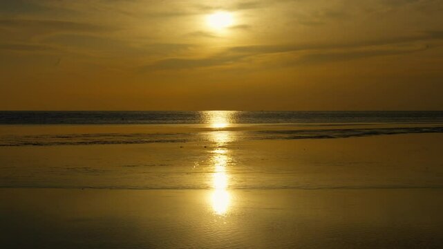 A tranquil beach scene at sunset features golden sunlight glimmering across the ocean&mdash;gentle cloud movement, subtle waves, and the reflective wet sand create a quiet coastal moment of natural harmony.