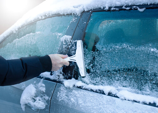 Man scraping ice from car window during snowy winter day. Concept of winter driving, vehicle maintenance, cold weather and road safety.