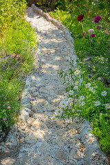 Beautiful handmade stone path in a blooming garden. The walkway is surrounded by flowers  daisies, lavender, and Turkish carnations. Natural landscape design, cozy countryside atmosphere, summer day, 