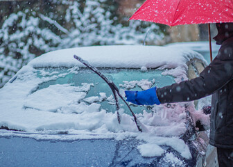 Person cleaning snow from car windshield during snowfall while holding a red umbrella. Winter...