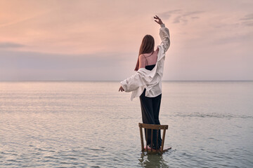 Young woman stands gracefully on chair in calm water, framed by serene sunset. Her pose evokes...