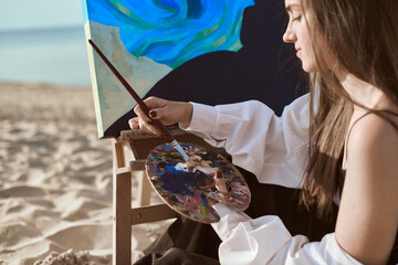 Young woman artist paints by sea, blending colors on palette. Her serene expression reflects...