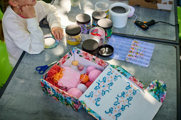 Child with light skin focuses on yarn craft at outdoor table. Bright yarn, organized supplies, and floral-patterned box enhance vibrant, creative setting
