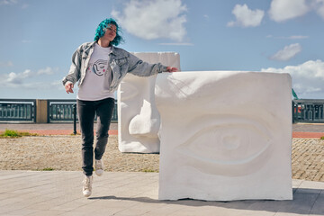 Young adult woman with blue hair and denim jacket walking near large white abstract sculptures in outdoor setting. Sunny day with clear sky enhances vibrant scene
