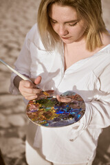 Young woman artist stands on sandy beach, engrossed in her painting. She holds colorful palette and...