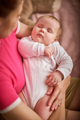 Young woman with braided hair gently cradles sleeping baby girl in pink and beige room. Warm lighting and soft textures create calm atmosphere