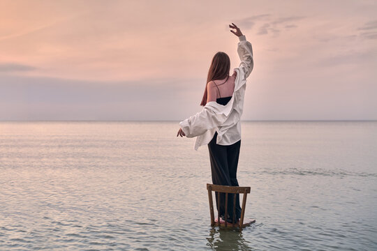 Young woman stands gracefully on chair in calm water, framed by serene sunset. Her pose evokes sense of balance and freedom, blending with tranquil seascape