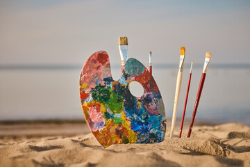 Vibrant painter palette and brushes rest on tranquil beach, symbolizing creativity and inspiration....