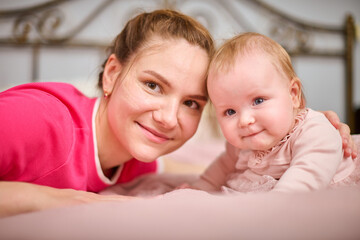 Young mother with brown hair, smiling baby girl indoors. Warm pink tones, soft lighting, metal bedframe in background. Expression of joy and connection