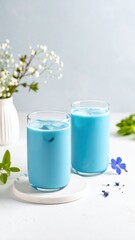 Refreshing blue beverages served in two transparent glasses with ice, surrounded by greenery and delicate flowers on a light background