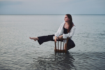 Young woman sits on chair in calm sea waters at dusk, capturing balance and tranquility. Her...