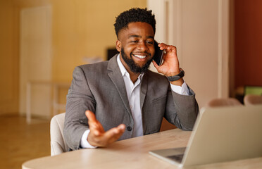 Businessman smiling and discussing with customer on phone call while using laptop at desk in office