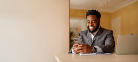 Portrait of confident businessman with laptop on desk messaging over mobile phone against wall in modern office