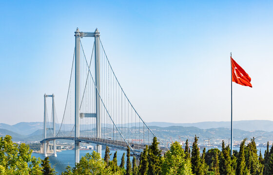 A majestic suspension bridge, the Osmangazi Bridge, spans a vast body of water, with vehicles crossing under a clear blue sky. A prominent Turkish flag proudly flies in the foreground.