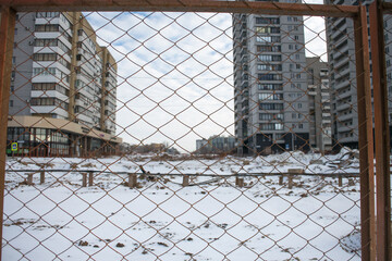 Snow covers a vacant construction site amid tall, modern buildings. A chain-link fence encloses the area, showcasing an incomplete urban project in a chilly environment.