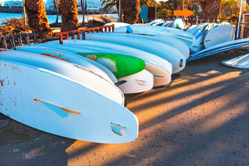 Colorful paddleboards and surfboards rest upside down on a sunlit promenade near the beach, casting...
