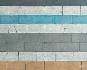 A vibrant, close-up overhead shot of a paving stone pattern featuring rectangular blocks in grey, white, and brown, accented by a bold blue stripe. Showcases rough textures and geometric forms.