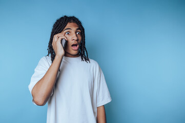 young man with dreadlocks talks on a smartphone, displaying astonishment and surprise on blue...