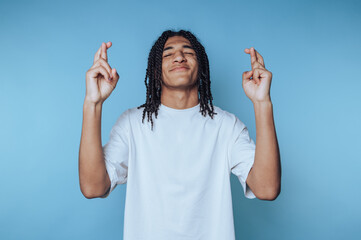 young man with dreadlocks stands with fingers crossed, displaying hope and anticipation on blue...