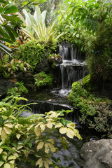 Old stone architecture of a Japanese garden with a fountain and pond, surrounded by trees, plants, and summer flowers