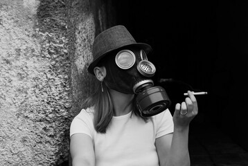 A woman in a gas mask and hat holds a cigarette while leaning against a textured wall
