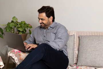 Fototapeta premium Indian handsome smiling man taking on phone while relaxing using laptop computer working and video conference meeting at work from home