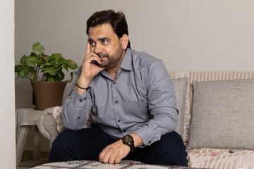 Indian unhappy sad man sitting on sofa alone at home, suffering from depression, loneliness or anxiety, copy space. Permacrisis, midlife crisis, financial troubles, lack of money and health problems