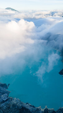 Aerial view of a turquoise lake nestled in a volcanic crater, shrouded in ethereal clouds, with rugged rock formations in the foreground, Ijen Volcano Crater, East Java, Indonesia.