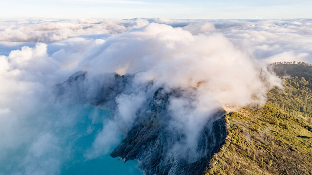 Aerial view of the rugged volcano crater veiled in ethereal white clouds, contrasting sharply with the turquoise lake and verdant slopes, Ijen Volcano Crater, East Java, Indonesia.