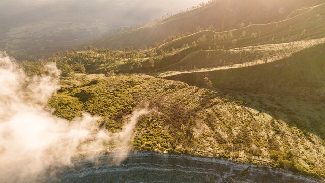 Aerial view of mist-shrouded slopes and terraced hillsides meet rugged cliffs under a soft, diffused light, Ijen Volcano Crater, East Java, Indonesia.