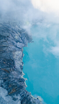 Aerial view of the turquoise lake nestled in the crater, bordered by rugged cliffs shrouded in ethereal mist, Ijen Volcano Crater, East Java, Indonesia.