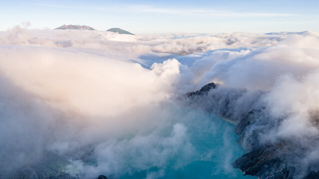 Aerial view of the turquoise crater lake embraced by swirling clouds, with distant peaks piercing through the misty veil, Ijen Volcano Crater, East Java, Indonesia.