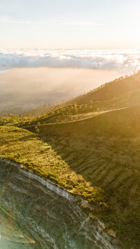 Aerial view of the sun-kissed, rugged terrain meeting the soft, ethereal clouds, a dance of light and shadow upon Ijen Volcano Crater, East Java, Indonesia.