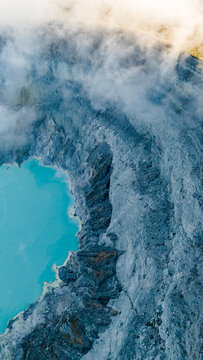 Aerial view of the breathtaking contrast of the vibrant turquoise lake against the rugged, textured volcanic crater, shrouded in ethereal white smoke, Ijen Volcano Crater, East Java, Indonesia.